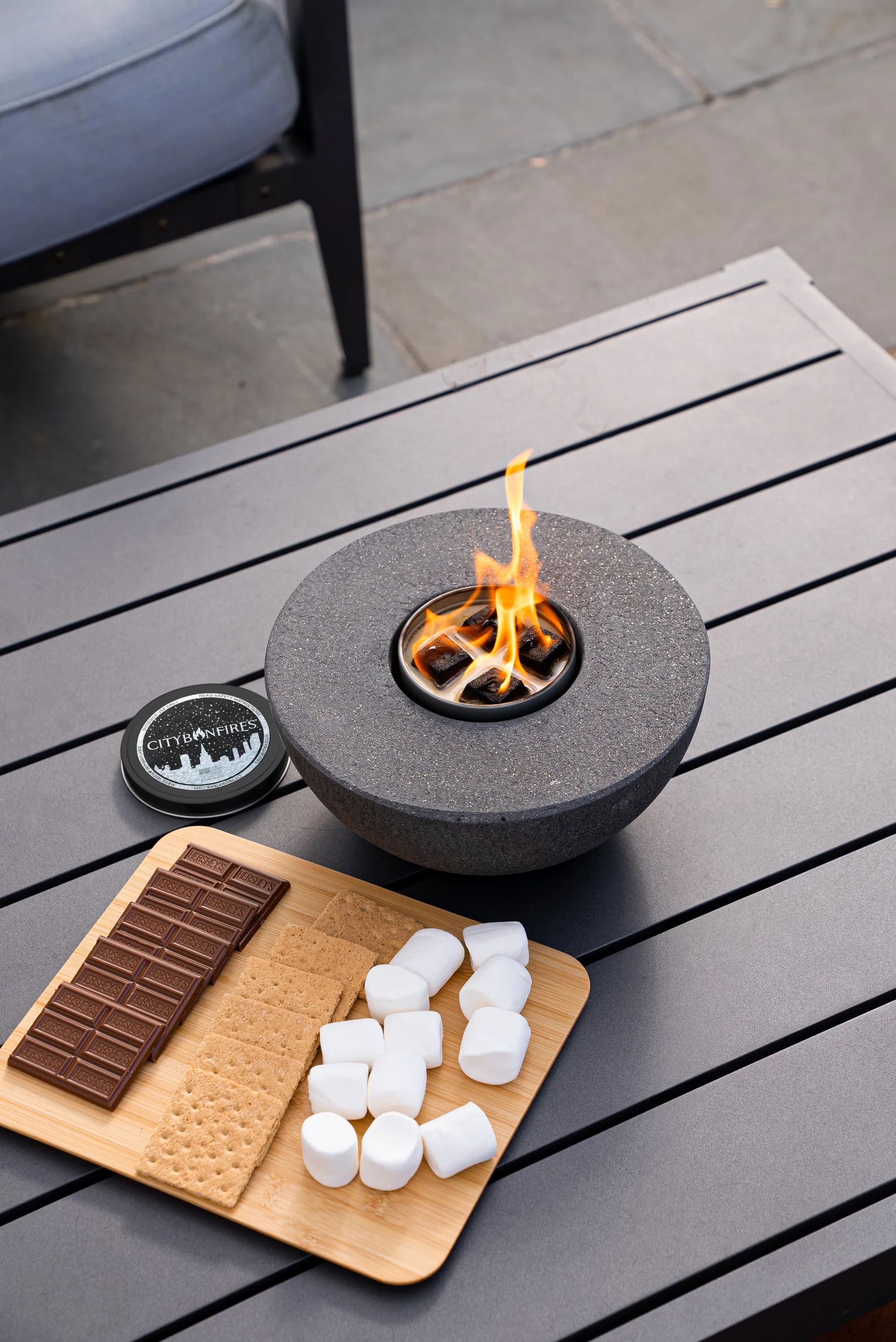 Lit concrete fire pit with marshmallows and chocolate on a table, city lights blurred behind